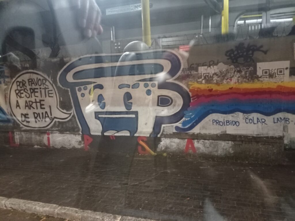 Foto de um graffiti num muro, com as frases "por favor respeite a arte de rua!" e "proibido colar lamb". O desenho do graffiti é de uma caneca com um rosto sorridente, do artista "Cabeça de xícara"