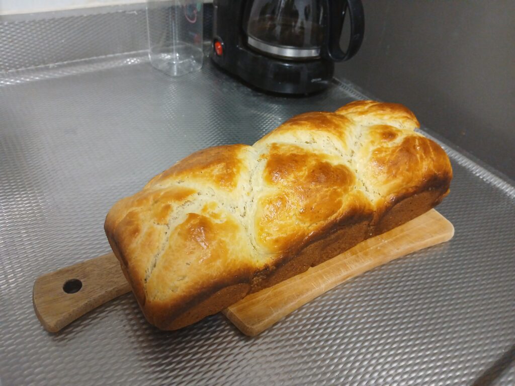 Foto de um pão do tipo brioche, em cima de uma tábua de madeira. No fundo tem uma cafeteira.