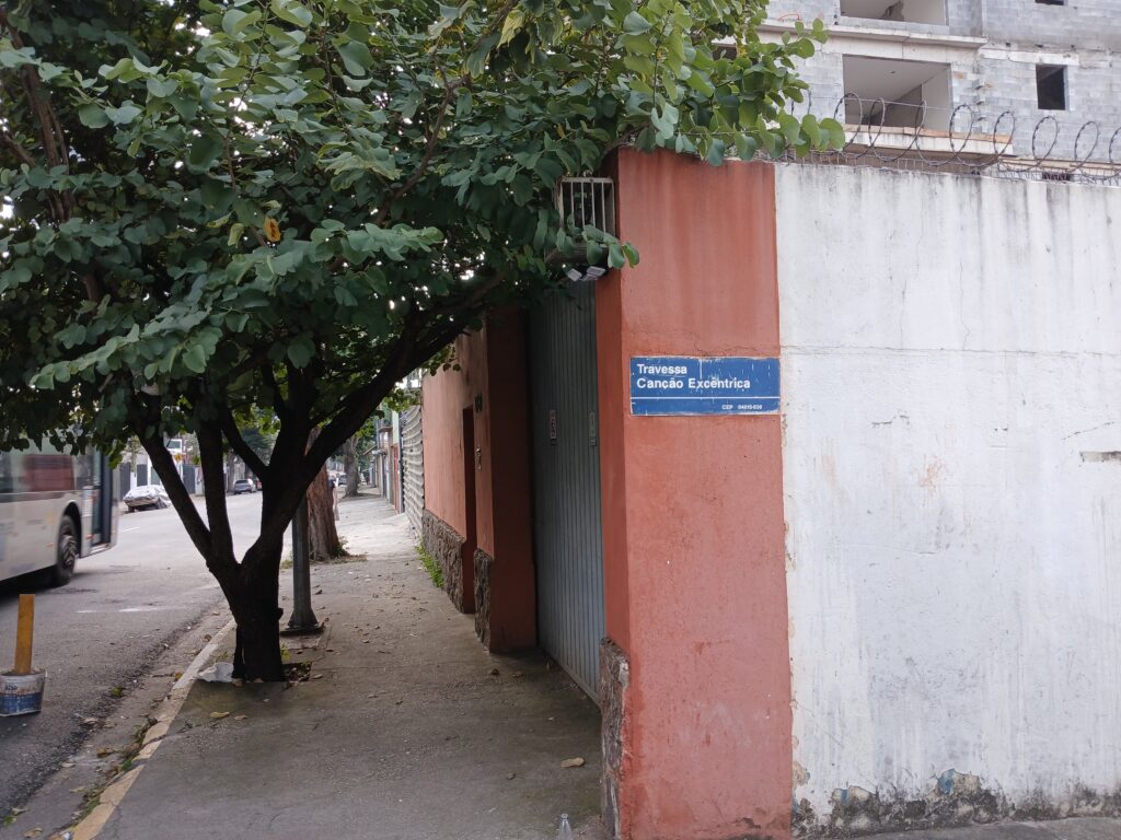 Foto de uma entrada de uma travessa na rua. À esquerda é possível ver um pedaço da rua, uma calçada e umas árvores. À direita, um muro com uma placa descrevendo o nome da travessa. Na placa diz "Travessa Canção Excêntrica".