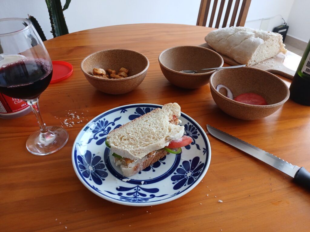 Foto de uma mesa de madeira. Na foto podemos ver um prato com um sanduíche, uma taça de vinho tinto, três potes com tomate, frango refogado e pimentão, e um pão ao fundo.