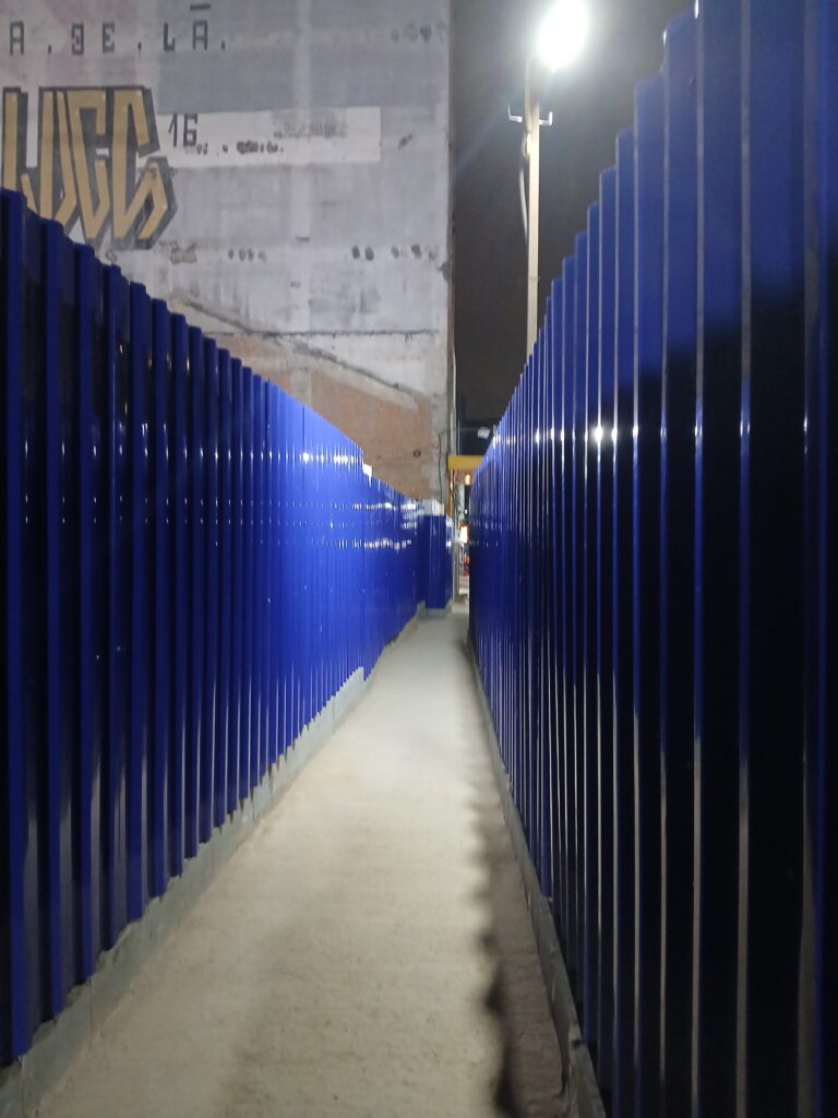 Foto de um corredor com paredes metálicas azuis, para proteger as pessoas enquanto passam perto de uma obra. No fundo é possível ver um prédio antigo com a pintura desbotada.