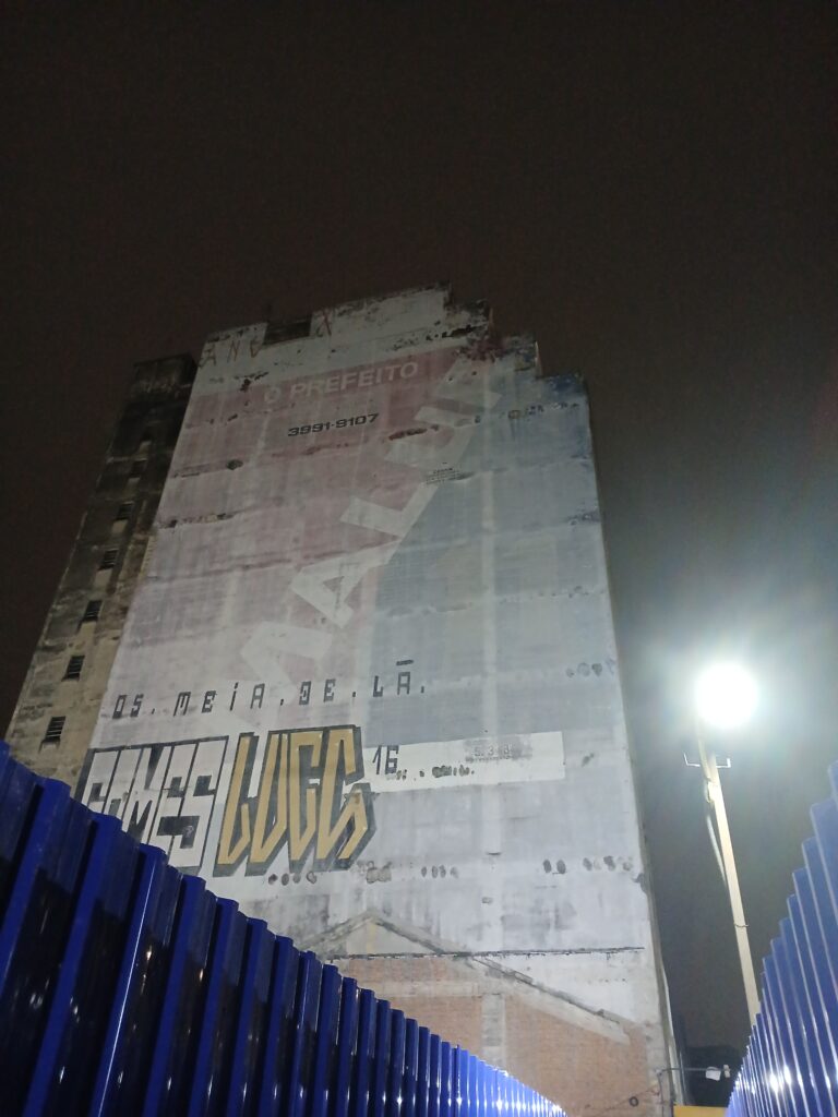Foto tirada à noite, mostrando a lateral de um prédio antigo. Na parede tem alguns grafitis e outras pinturas desbotadas.