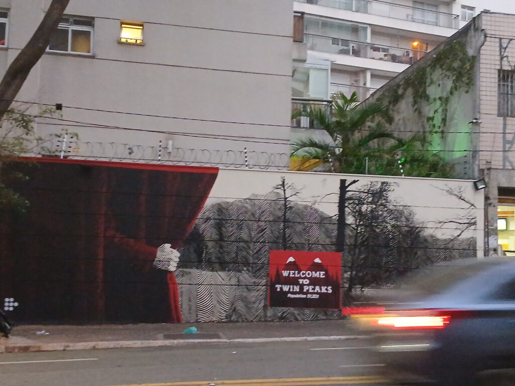 Foto tirada na rua Augusta, mostrando um muro com uma propaganda da série Twin Peaks. O desenho mostra uma floresta em preto e branco, e uma placa vermelha com a frase "welcome to Twin Peaks". Na frente da foto, tem um carro passando.