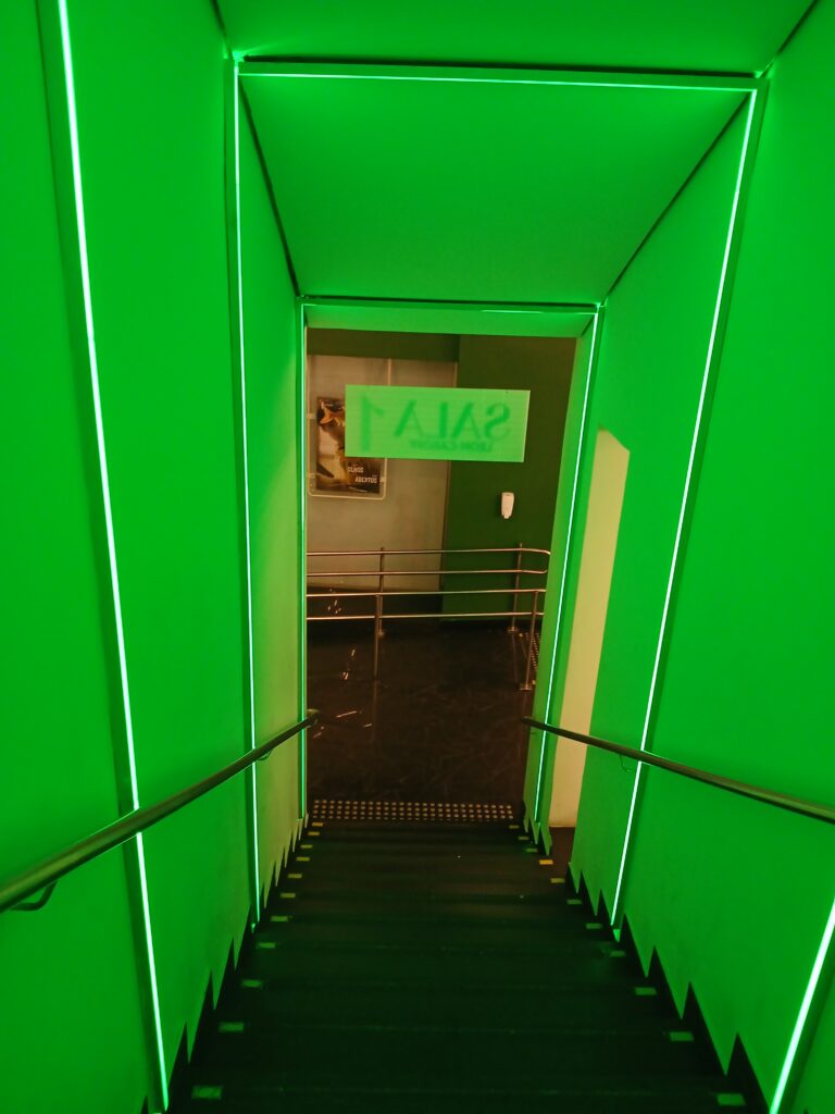 Foto de uma escadaria em um corredor, visto de cima, do ponto de vista de quem vai descer. As paredes e o teto são brancos, mas ficam totalmente verdes por causa da luz verde. Na entrada, tem uma placa escrito "sala 1", mas como está virada de costas para quem desce a escada, aparece escrito invertido na horizontal.