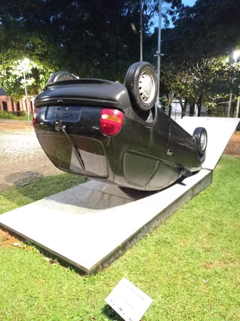Foto da instalação chamada "cena de cinema" de José Resende. A obra é um carro Celta preto capotado, ou seja, virado com as rodas pra cima. A obra fica num gramado verde