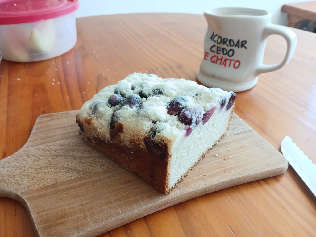 Foto de uma cuca de uva, sobre uma tábua de madeira. Ao fundo, à direita tem uma caneca de café branca com a frase "acordar cedo é chato", e à esquerda, um pote com tampa vermelha.