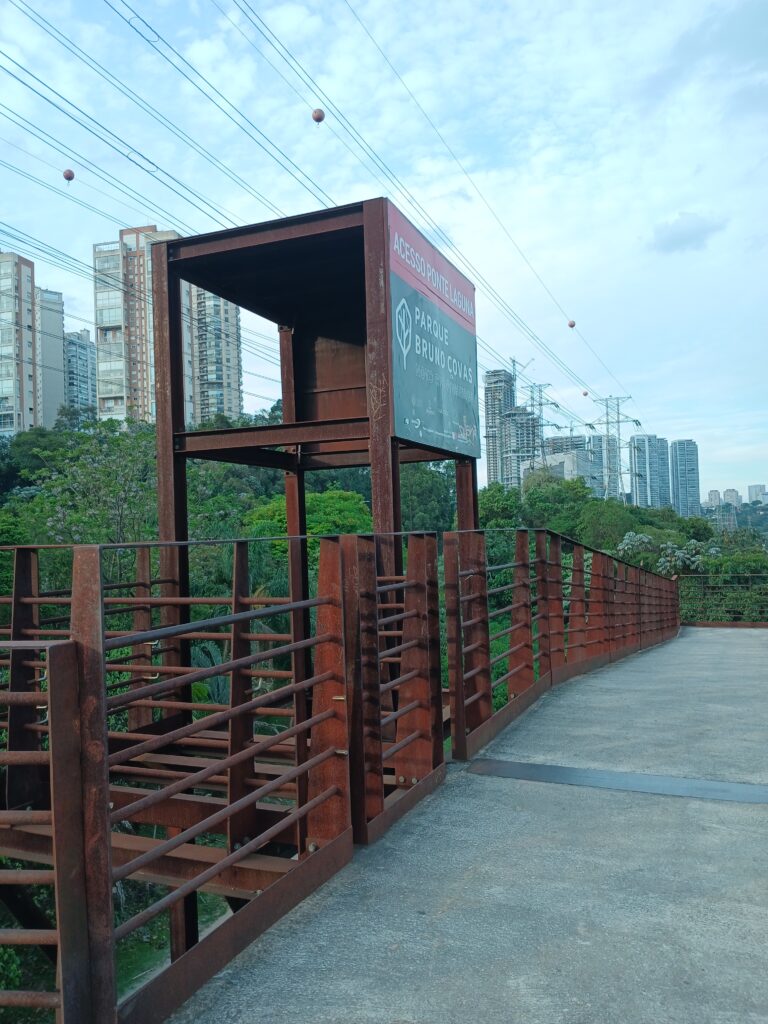 Foto mostrando o acesso da ponte Laguna ao parque. A foto mostra uma estrutura de metal enferrujada, com uma placa indicando o nome do acesso. Ao fundo tem algumas árvores, cabos e uma torre de transmissão de energia, e um monte de prédios.