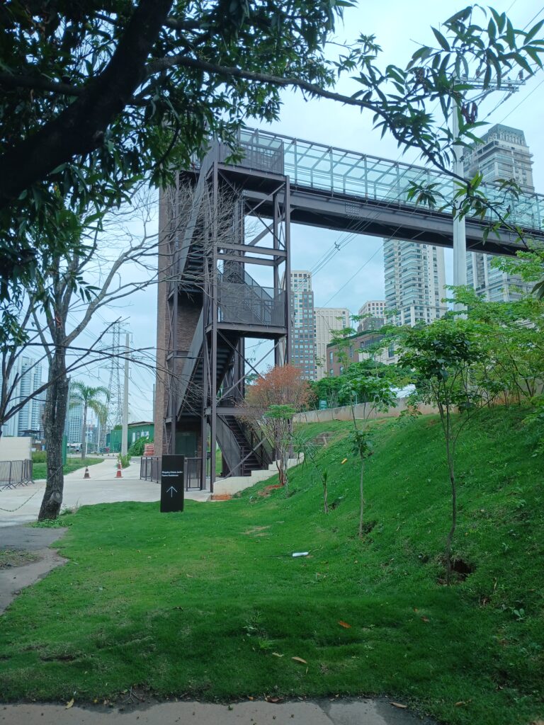 A foto mostra uma passarela, e uma escadaria e elevador que levam até a passarela. Há também algumas árvores e prédios ao fundo.