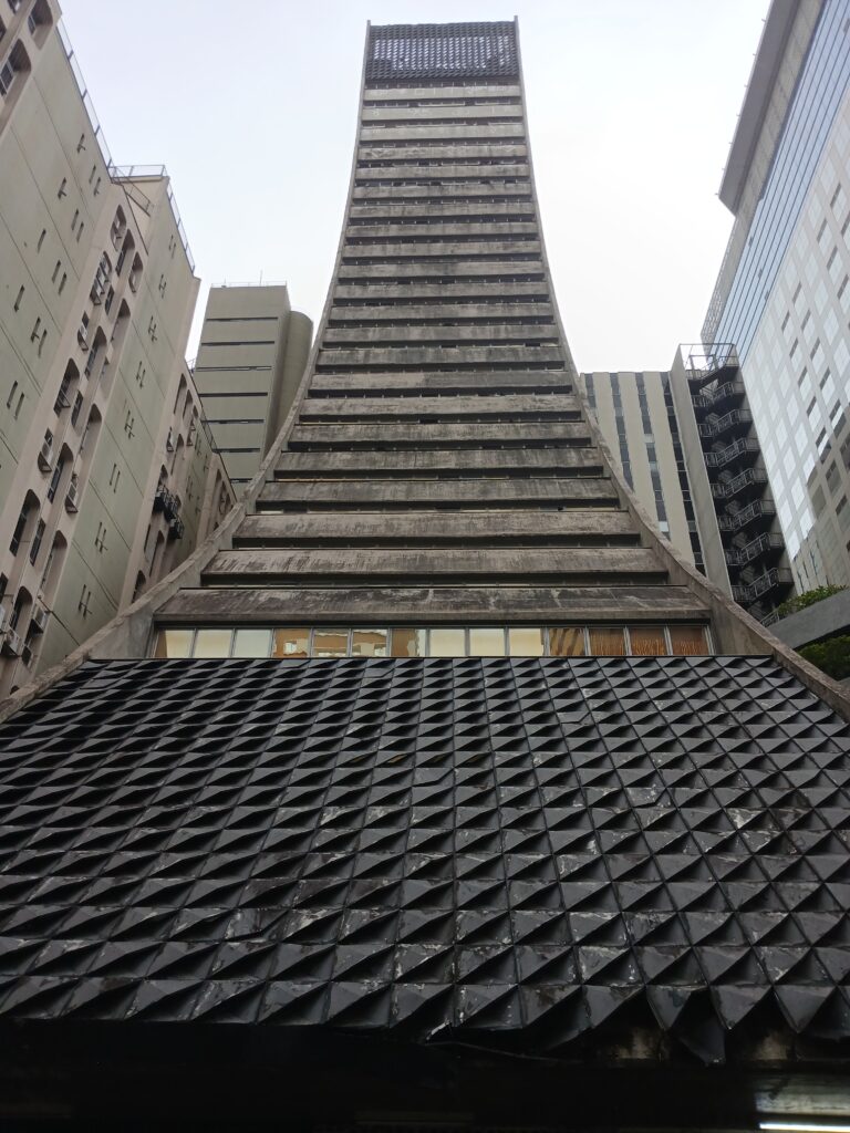 Foto de um edifício visto de baixo para cima. O formato diferente do prédio, que é como se fosse um funil invertido, torna a vista à partir desse ponto bem única.