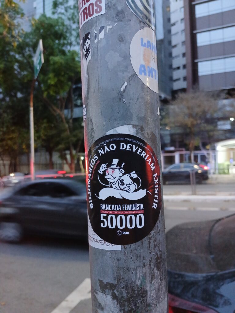 Foto de um poste mostrando um adesivo da bancada feminista do PSOL com a frase "bilionários não deveriam existir". Ao fundo, uma rua e alguns carros passando.