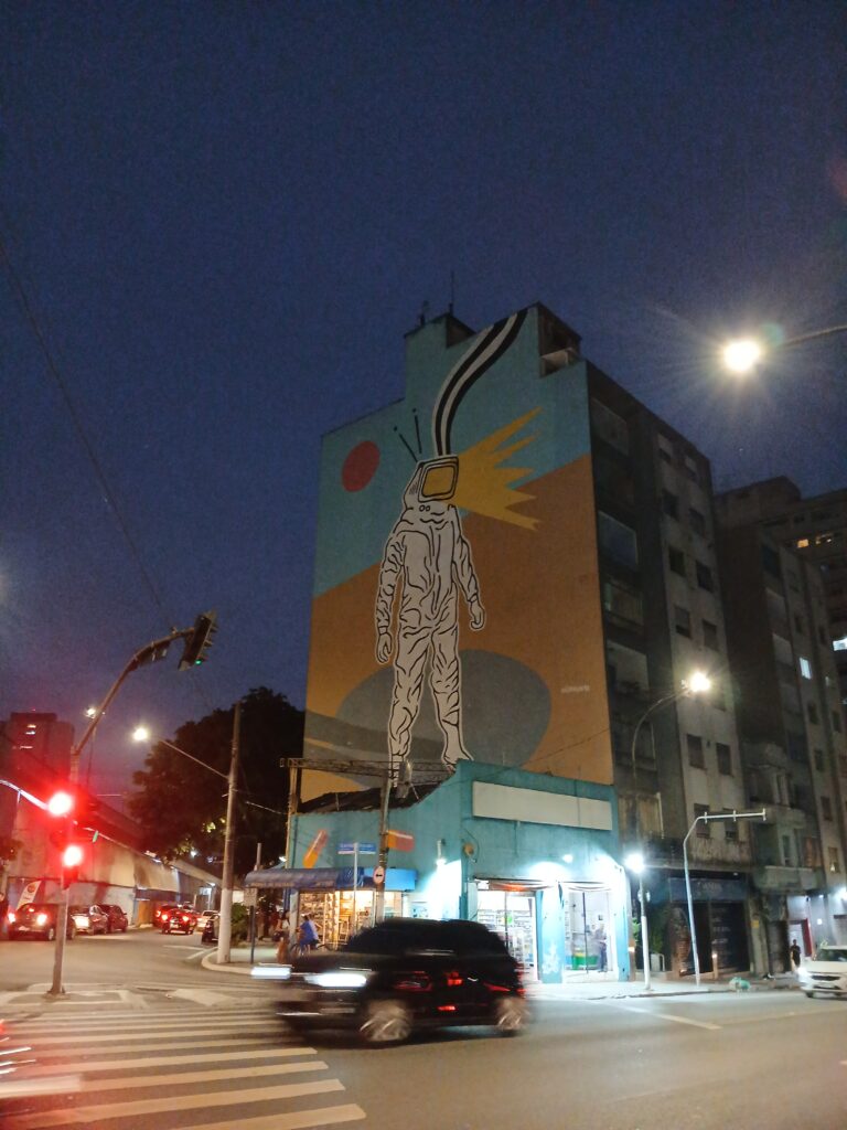 Foto de um prédio visto meio de lado, à noite. Na lateral do prédio tem um desenho gigante de uma pessoa em uma roupa branca parecendo de astronauta. No lugar da viseira do capacete tem uma tela de tv emitindo uma luz amarela, e ele tem duas antenas pequenas. Mais abaixo na foto é possível ver um carro passando e um comércio aberto.